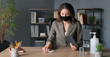 Maskeli güzel beyaz kadın ofis çalışanı masada oturuyor ve tablet cihazını peçeteyle siliyor. Coronavirus salgını koruması. Kadın temizliği, sterilize etme ve bilgisayarı sterilize etme.