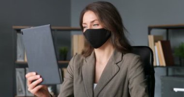 Maskeli şık, beyaz bir kadın ofisinde otururken tablet bilgisayarında video kaset taşıyor. Genç iş kadını web kamerasıyla konuşuyor ve internette görüntülü sohbet yapıyor. Kadın video blogcusu.