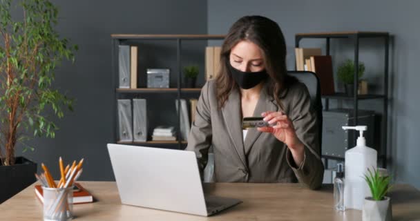 Young Caucasian female sitting at table in office with credit card in hand and shopping online on laptop computer. Pretty businesswoman in mask buying in internet and paying. Shopper on quarantine. — Stock Video