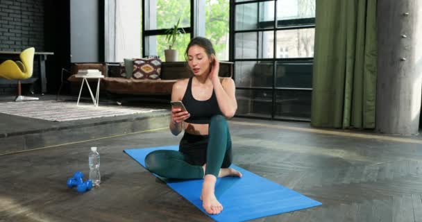 La joven caucásica guapa que se sienta en el tatami en el suelo y utiliza el teléfono inteligente para ver el ejercicio de fitness en línea en casa. Autoaislamiento cuarentena. Bandejas deportivas de niñas y niños en el teléfono móvil. — Vídeo de stock