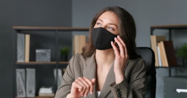 Caucasian young businesswoman sitting and talking on cellphone. Beautiful female office worker in mask speaking on mobile phone in cabinet. Coronavirus pandemic time. Telephone call. — Stock Video