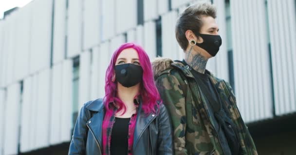 Portret białych hipsterek w czarnych maskach, stojących na ulicy od tyłu do tyłu. Stylowa młoda dziewczyna z różowymi włosami i facet z tatuażami na zewnątrz podczas koronawirusu. — Wideo stockowe