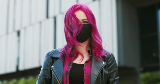 Ritratto di giovane ragazza caucasica piuttosto elegante con i capelli rosa indossando maschera nera e guardando la fotocamera all'aperto. Primo piano di donna straordinaria con colore lucente di capelli in protezione respiratoria . — Video Stock