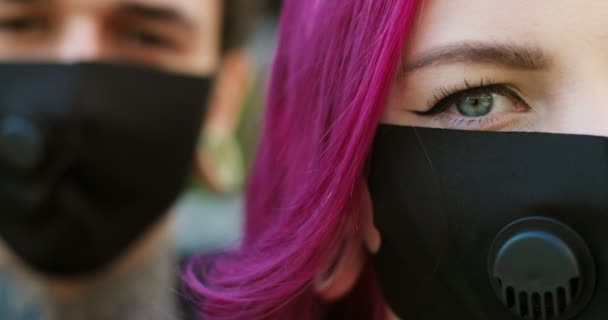 Close-up de metade das faces de jovens hipsters caucasianos em máscaras respiratórias pretas olhando para a câmera ao ar livre. Retrato de menina extraordinária com cabelo rosa e namorado na rua durante coronavírus . — Vídeo de Stock