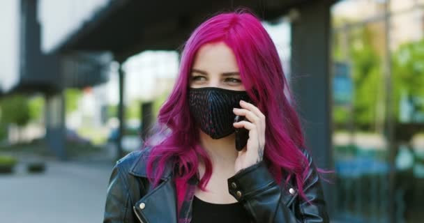 Καυκάσιος νεαρός κομψό κορίτσι με ροζ μαλλιά και μάσκα μιλάμε στο κινητό τηλέφωνο εξωτερική. Γυναίκα hipster μιλώντας στο κινητό στο δρόμο κατά τη διάρκεια της πανδημίας coronavirus. — Αρχείο Βίντεο