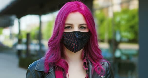 Портрет молодої красивої кавказької дівчини з рожевим волоссям у дихальній масці, дивлячись на камеру на вулиці. Покриття незвичайної самиці з яскравим кольором волосся в захисті від косид-19. — стокове відео