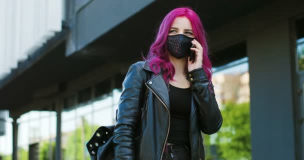 Happy, pembe saçlı ve maskeli, dışarıda cep telefonuyla konuşan, beyaz, fevkalade bir kız gibi gülümsedi. Koronavirüs salgını sırasında caddede cep telefonuyla konuşan neşeli bayan hipster.. — Stok video