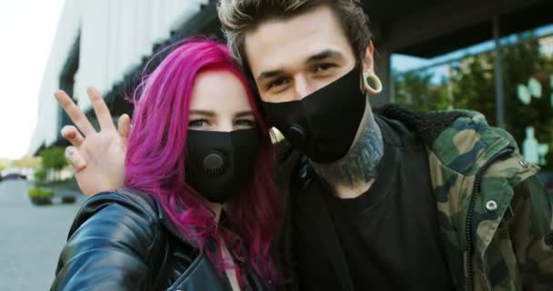 POV de chico elegante caucásico con tatuajes y chica con el pelo rosa hablando y agitando las manos a la cámara al aire libre durante la pandemia coronavirus. Extraordinaria pareja de hipsters en máscaras teniendo videochat . — Vídeo de stock