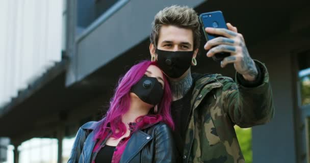 Caucásico pareja extraordinaria de hipsters en máscaras respiratorias de pie en la calle y tomar fotos selfie con la cámara del teléfono inteligente. Chica con estilo con el pelo rosa y chico al aire libre haciendo foto con el teléfono . — Vídeo de stock