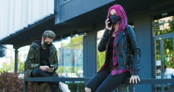 Caucasian couple of extraordinary hipsters in masks keeping social distance outdoor. Girl with pink hair talking on mobile phone and guy texting or scrolling on smartphone at street. Streeters. — Stock Video