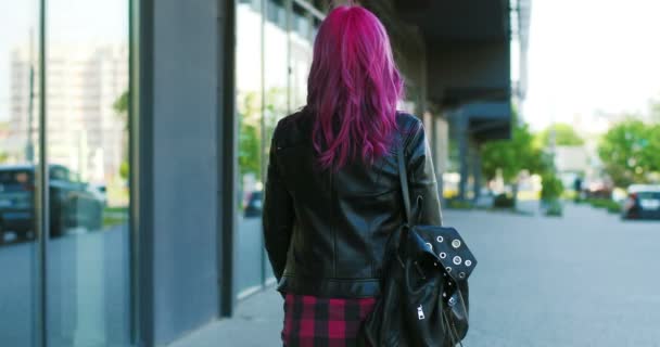 Задній бік молодої кавказької екстраординарної дівчини з рожевим волоссям і в чорній шкіряній куртці, що йде на вулицю, а потім звертається до камери. На задній погляд на стильний жіночий гіпстер прогулюючись вулицею і повертаючись. — стокове відео