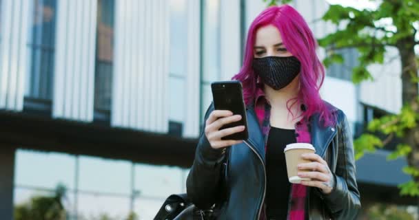 Jeune fille caucasienne extraordinaire avec les cheveux roses et dans le masque debout à l'extérieur, avec café à emporter et message texte sur smartphone. Élégant défilement hipster féminin et tapoter sur le téléphone mobile . — Video