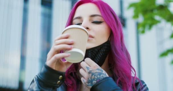 Primer plano de la joven chica extraordinaria caucásica con el pelo rosa y tatuaje de pie al aire libre y quitándose la máscara para beber café para llevar. Elegante hipster femenino sorbiendo té caliente. Beber en la calle . — Vídeo de stock