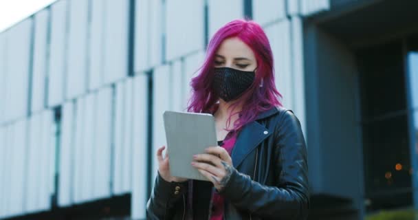 Νεαρό Καυκάσιο εξαιρετικό κορίτσι με ροζ μαλλιά και μάσκα στέκεται σε εξωτερικό χώρο και γραπτών μηνυμάτων στη συσκευή tablet. Κομψό γυναικείο hipster κύλιση, δακτυλογράφηση και πάτημα στον υπολογιστή σε απευθείας σύνδεση. — Αρχείο Βίντεο