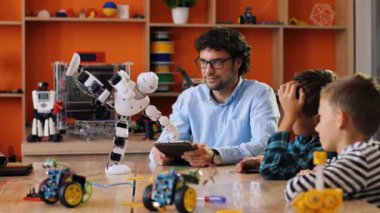 Yakışıklı bir adam çocuklara oyun alanındaki tablet cihazıyla robot oyuncağı kullanmayı öğretiyor..