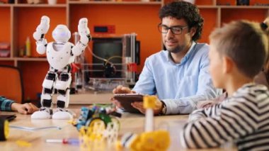 Gözlüklü yakışıklı öğretmen çocuklara tablet cihazını kullanarak robotu nasıl kullanacaklarını ve modern oyun odasında nasıl hareket ettireceklerini gösteriyor. Kapalı
