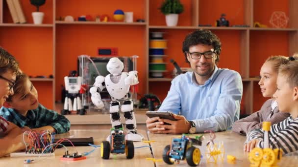 Profesor masculino en las gafas con los niños viendo el robot moviéndose divertido en la mesa en el salón de clases moderno y colorido. De interior — Vídeos de Stock