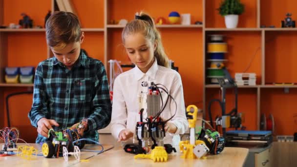 Portrait shot of schoolgirl and schoolboy putting together robots in front of the camera and smiling in the nice classroom — Stock Video