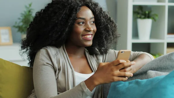 Mooie krullende Afro-Amerikaanse jonge vrouw chatten en lachen op haar smartphone. Woonkamer. Portret — Stockfoto