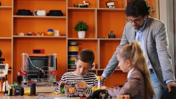 Guru tampan pria mengendalikan bagaimana anak laki-laki dan perempuan merakit robot di kelas modern yang diperlengkapi dengan baik. Guru puas dengan hasilnya. — Stok Video