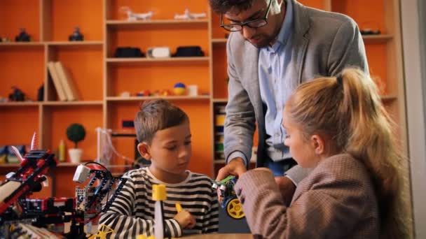 Male teacher controlling and helping to children in assembling robots in good equipmented modern classroom space. — Stock Video