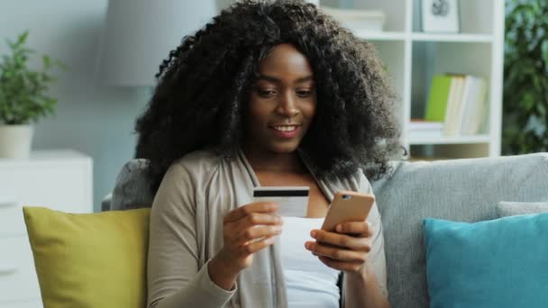 Joven bastante afroamericana mujer compras en línea con tarjeta de crédito utilizando su teléfono inteligente mientras está sentado en el sofá. De interior . — Vídeo de stock