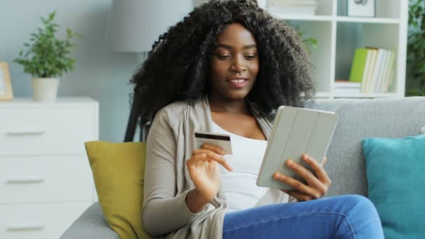 Ganska afroamerikansk ung kvinna online shopping med kreditkort med hjälp av hennes vita tablett enhet. Inomhus. — Stockvideo