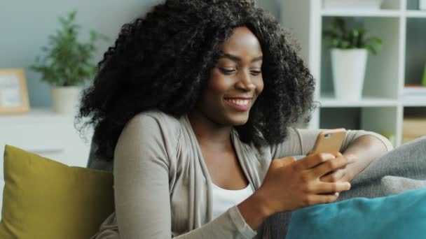 Schöne lockige afroamerikanische junge Frau, die mit ihrem Smartphone plaudert und lacht. Wohnzimmer. Porträt — Stockvideo