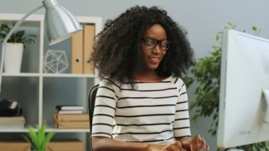 Tatmin olmuş, gülümseyen, gözlüklü, bilgisayar başında çalışan ve ofiste rahatlayan kıvırcık saçlı, Afro-Amerikan kadın. Kapalı