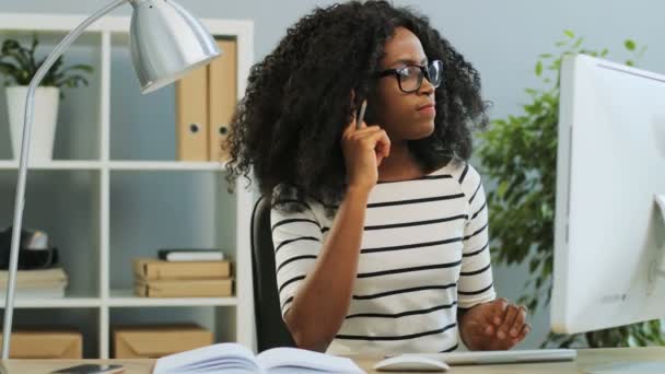 Ziemlich lockige junge Afroamerikanerin mit Brille, die am Personal Computer arbeitet und im Büro Notizen im Notizbuch macht. — Stockvideo