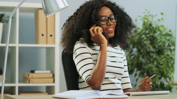 Retrato de la hermosa rizada joven afroamericana en gafas que hablan en el teléfono móvil en la oficina. De interior — Vídeo de stock