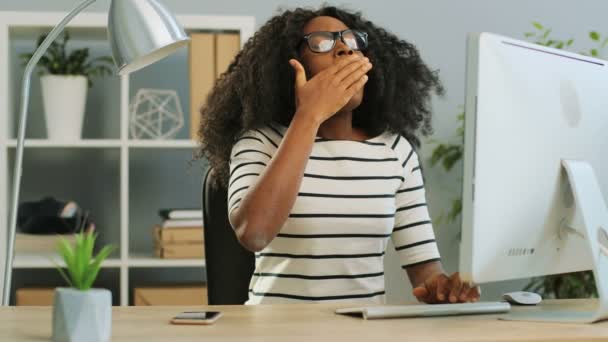 疲れと眠い若いアフリカ系アメリカ人の女性がオフィスでコンピュータに取り組んでいる間、眼鏡をかけてあくびをする. — ストック動画
