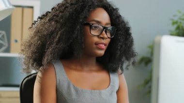 Bilgisayarda çalışan ve ofiste düşünen, gözlüklü, güzel, kıvırcık, Afro-Amerikan kadın. Portre çekimi