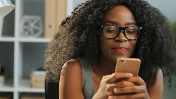 Close-up van mooie krullend glimlachte Afro-Amerikaanse jonge vrouw sms 'en op haar smartphone in het kantoor. Portretfoto. Binnenshuis — Stockvideo