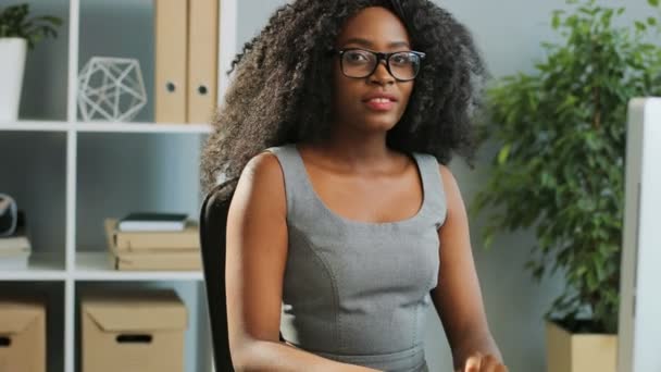 Großaufnahme der schönen lockigen jungen Afroamerikanerin mit Brille, die im Büro am Computer arbeitet und lächelt. Porträtaufnahme. Innenräume — Stockvideo