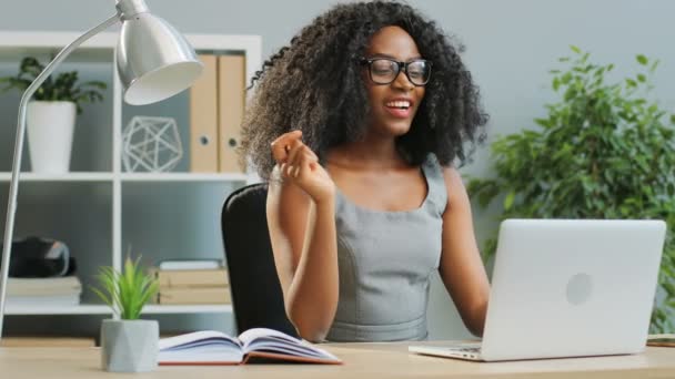 Rizado joven bonita mujer afroamericana en gafas que tienen video chat en la computadora portátil en la oficina y agitando su mano. De interior — Vídeos de Stock