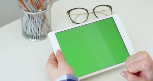 Белое планшетное устройство с зеленым экраном в руках белой женщины за столом. Женщина прокручивает, масштабирует и нажимает на хроматический ключ экрана горизонтального компьютера за столом. Просмотр и серфинг онлайн . — стоковое видео