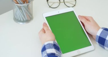 Masadaki beyaz tenli kadının ellerinde yeşil ekranlı beyaz tablet. Kadın kaydırma, yakınlaştırma ve masadaki bilgisayarda krom tuş ekranına tıklama. Çevrimiçi tarama.
