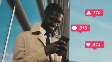 Genç, Afro-Amerikalı, gözlüklü yakışıklı blogcu iş merkezinde dikiliyor ve akıllı telefon kullanıyor. Sosyal medyada tepkiler, yorumlar, beğeni ve kalp alan adam.