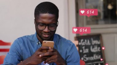 Gözlüklü Afro-Amerikalı genç erkek blogcu kafede oturuyor ve sosyal medyada paylaşıyor. Adamın yeni takipçileri ve kalpleri online oluyor. İnternetteki iletişim ve tepkiler.