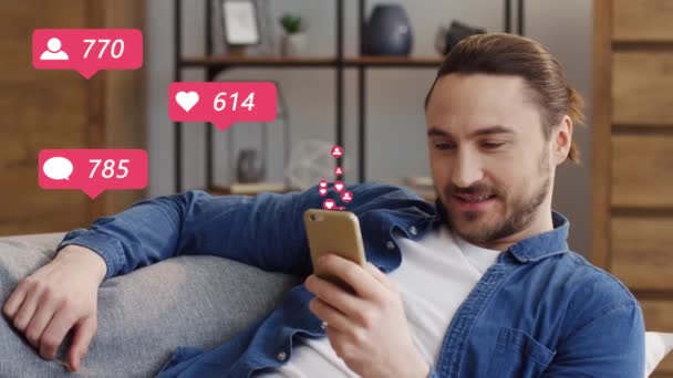 Handsome Caucasian young man sitting on couch in living room, watching posts and liking with hearts on smartphone. Male blogger using mobile phone and getting reactions and likes from followers. — Stock Video