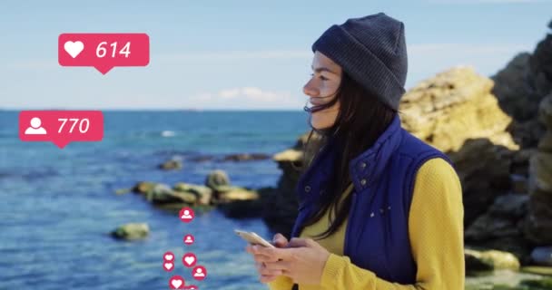 Stylish Caucasian young woman walking at rocky seacoast tapping and scrolling posts on smartphone. Female blogger at sea shore with phone getting reactions, likes and hearts from followers. — Stock Video