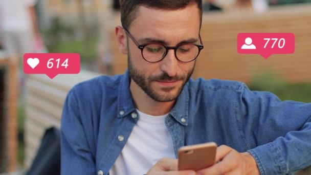 Νεαρός Καυκάσιος άνδρας με γυαλιά κάθεται σε υπαίθριο χώρο στην πόλη στο δρόμο και αναρτά μηνύματα στα μέσα κοινωνικής δικτύωσης στο smartphone. Guy blogger συγκέντρωση likes και οπαδούς σε απευθείας σύνδεση. Αρσενικό χρήστη αρέσει στο κινητό τηλέφωνο. — Αρχείο Βίντεο