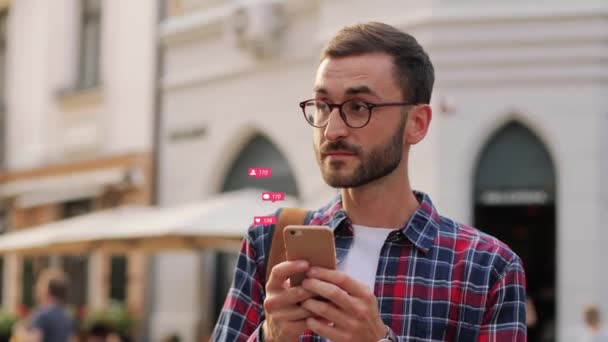 Pria muda berkacamata berdiri di luar kota di jalan dan mengirim pesan di media sosial melalui smartphone. Guy blogger mengumpulkan suka dan pengikut secara online. Pengguna laki-laki menyukai ponsel. — Stok Video