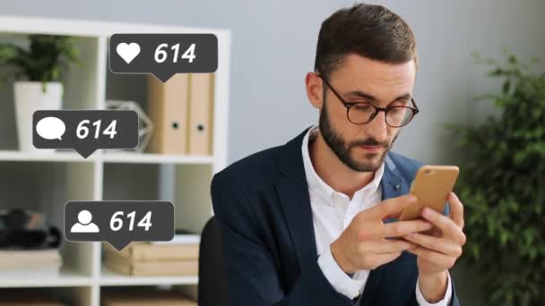 Gözlüklü yakışıklı, beyaz, genç bir işadamı ofisinde oturmuş cep telefonunda kalpleri olan yazıları izliyor. Erkek blogcu akıllı telefon kullanıyor ve takipçilere yorum ve gönderi için beğeni alıyor — Stok video