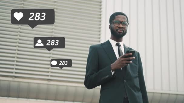 Afrikai amerikai fiatal jóképű üzletember öltönyben, nyakkendőben és szemüvegben kültéri hirdetési üzenőfal áll okostelefonon. Elérése sok szeret, megjegyzések és új követői a szociális média. — Stock videók