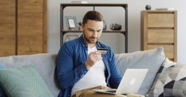 Pantolon gömlekli genç çekici bir adam elinde kredi kartıyla kanepede oturuyor ve rahat odadaki dizüstü bilgisayardan alışveriş yapıyor. İçeride.