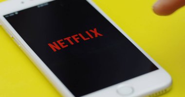 New York - 01 Aralık 2017: Netflix uygulamasını açan ve sarı masanın üzerinde duran beyaz akıllı telefonda kaydıran erkek parmağını kapat.