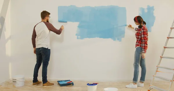 View from back on the Caucasian young woman in the plaid shirt and man in glasses painting a wall in the blue colour while doing repair at their apartment.. Indoors