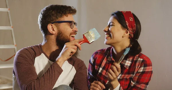 Close up of the young attractive funny man and woman painting on each others faces with big brushes for walls during a repair at their apartment. Indoor